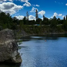 Kamienista plaża nad błękitną wodą, otoczona zielenią. Na wodzie bojki, w tle las i błękitne niebo z chmurami.