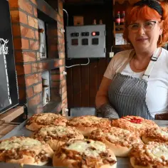 Kobieta z rudymi włosami prezentuje świeżo upieczone pizze. To może być najstarsza pizzeria w Polsce, z piecem opalanym drewnem w tle.