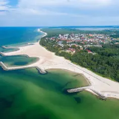 Widok z lotu ptaka na kładno nad morzem, z piaszczystą plażą, kamiennymi falochronami i zielonymi lasami otaczającymi miasteczko.