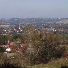 Widok na malowniczą wieś Gdów z kościołem i domami z czerwonymi dachami, otoczoną pagórkami i drzewami.