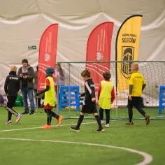 Dzieci trenują piłkę nożną w hali sportowej. W tle flagi "Akademia Piłkarska Sulechów", świadczące o wydarzeniach sportowych w Sulechowie.