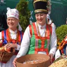 Trzy kobiety w strojach ludowych prezentują kosz jabłek i tacę z napojami podczas myszyniec wydarzenia.