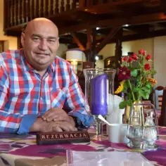 Łysy mężczyzna w kratę siedzi przy stole w karczmie. Na stole wazon z kwiatami, świeca i tabliczka "REZERWACJA".
