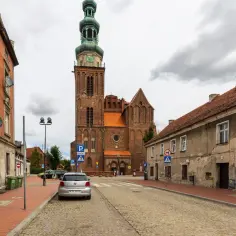 Ulica w Chełmży z widocznymi zabytkami: ceglanym kościołem z zieloną wieżą i kamienicami.