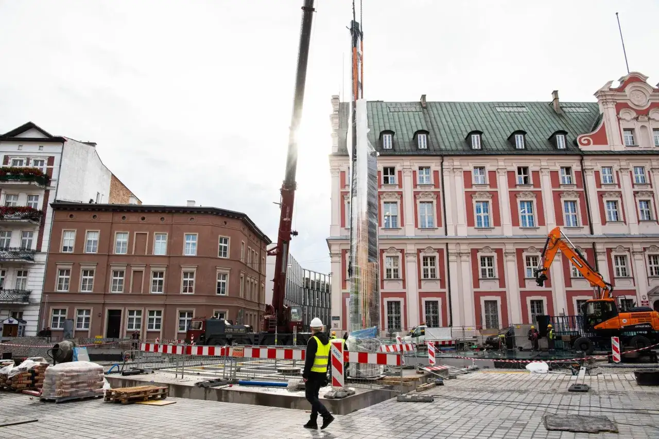 Plac Kolegiacki w Poznaniu podczas budowy. Dźwig podnosi lustrzaną instalację, obok pracują koparki i robotnicy.