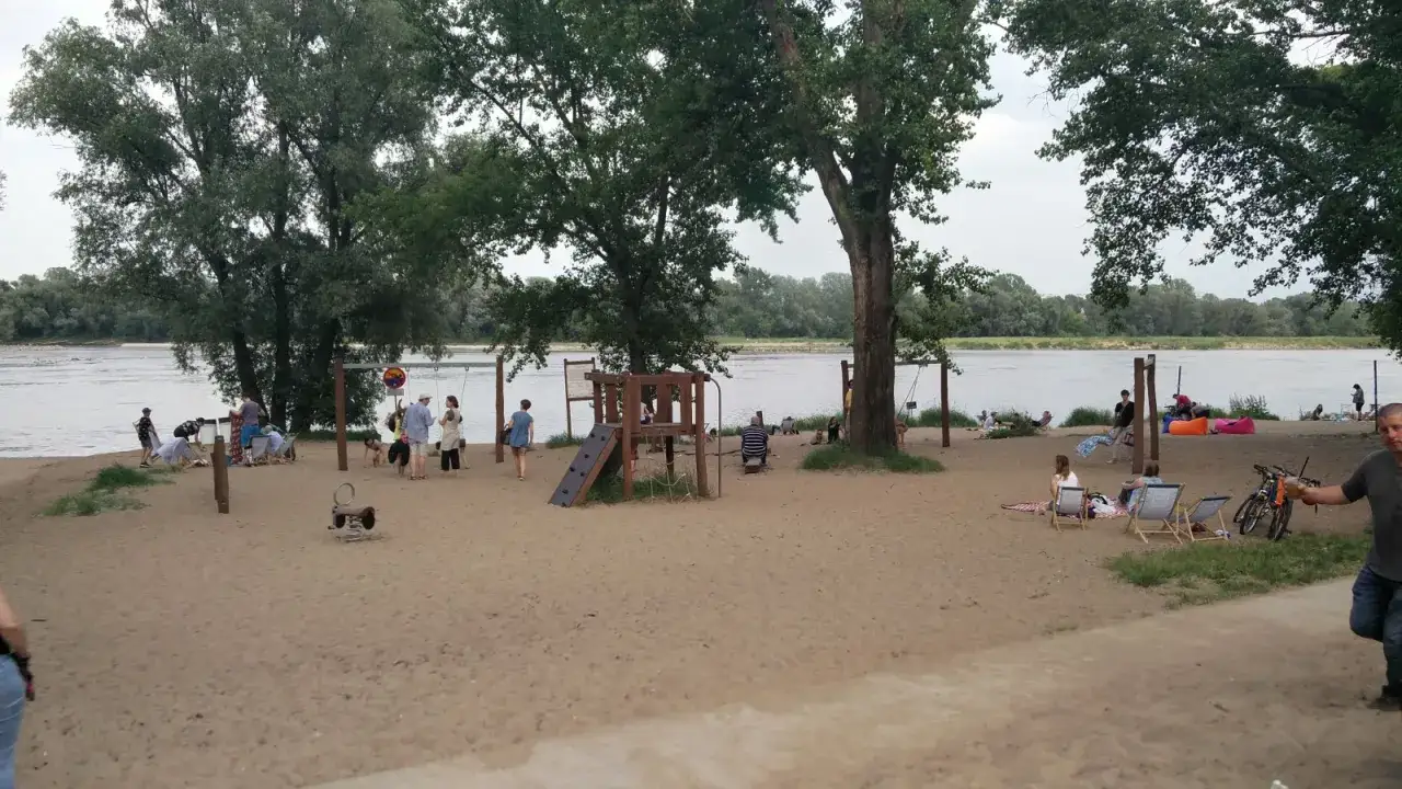 Plaża Żoliborz tętni życiem: dzieci bawią się na placu zabaw, dorośli odpoczywają na leżakach, a w tle widać Wisłę.