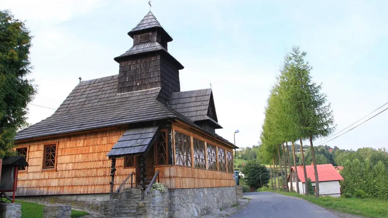 Drewniana cerkiew z wieżą, otoczona zielenią. W oddali domy z czerwonymi dachami. Laliki atrakcje zachwycają spokojem.
