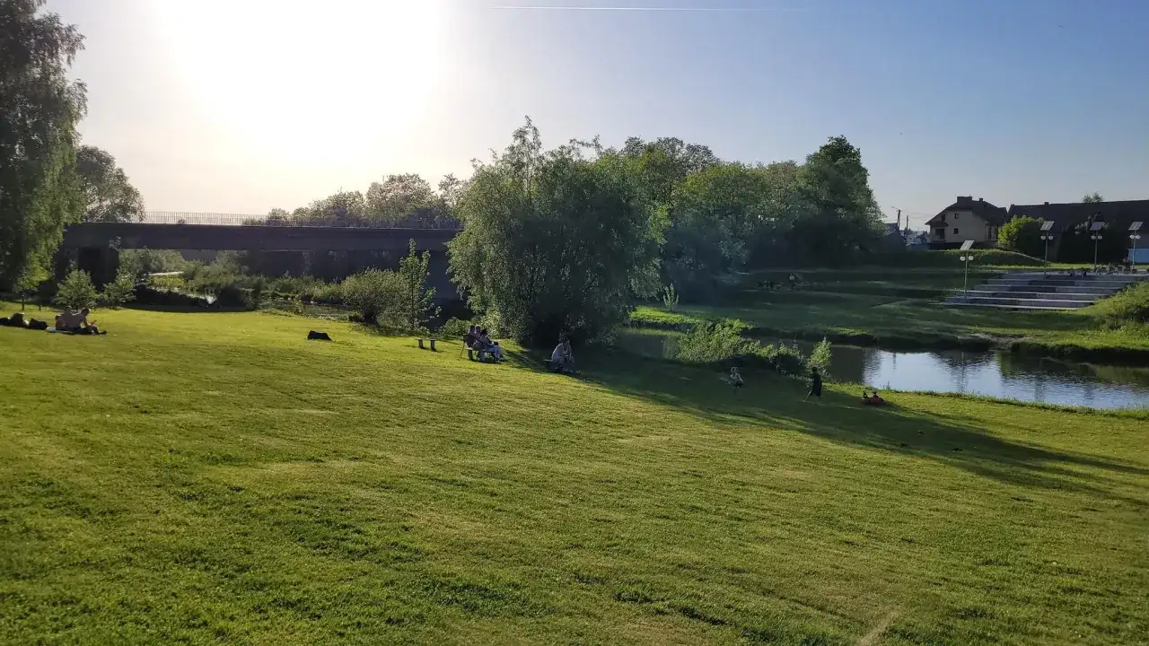 Słoneczny dzień nad rzeką w Gdowie. Ludzie odpoczywają na trawie, podziwiając widok na most i zieleń.
