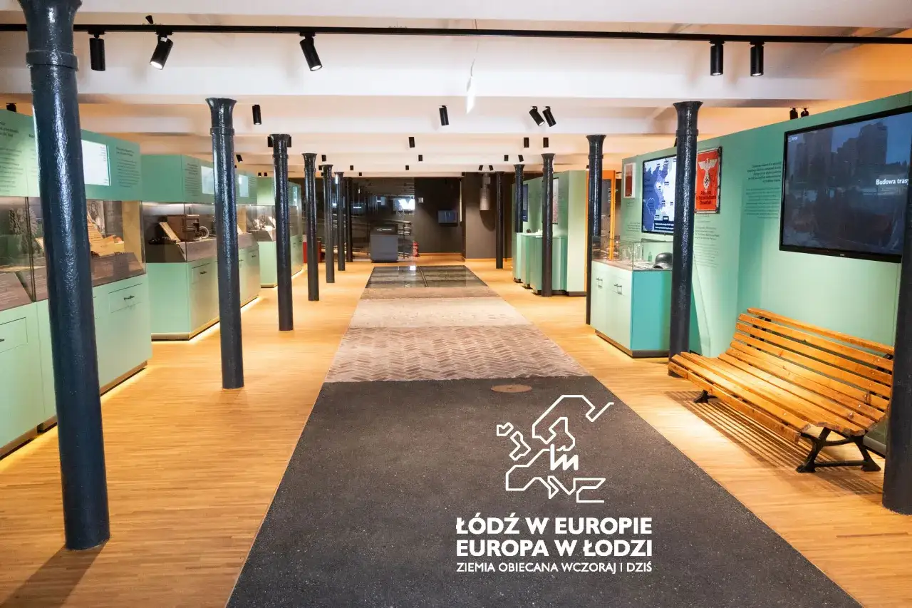 Wnętrze muzeum PRL w Łodzi. Ekspozycja z gablotami, ekranami i ławką. Na podłodze napis 