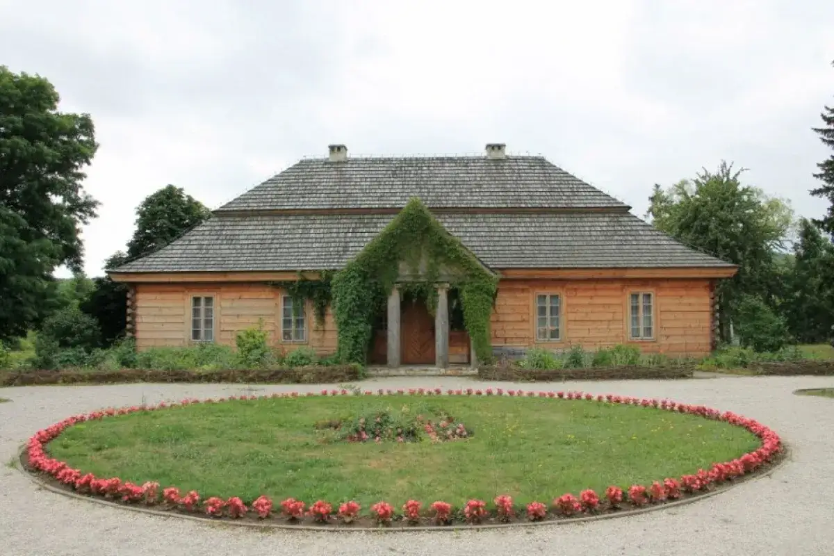 Drewniany dworek z werandą porośniętą bluszczem, otoczony ogrodem z klombem kwiatów. Ciekoty atrakcje czekają na zwiedzających!