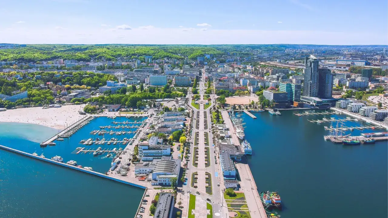 Widok z lotu ptaka na Gdynia Rynek Główny, z mariną pełną jachtów, plażą i zielonymi wzgórzami w tle.