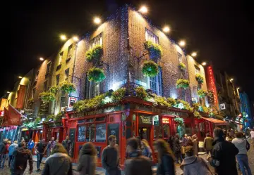 Nocne życie w Dublinie, irlandzka święta. Tłum ludzi spaceruje ulicą przed oświetlonym The Temple Bar, ozdobionym girlandami świateł i wiszącymi koszami z kwiatami.