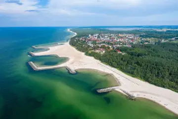 Widok z lotu ptaka na kładno nad morzem, z piaszczystą plażą, kamiennymi falochronami i zielonymi lasami otaczającymi miasteczko.