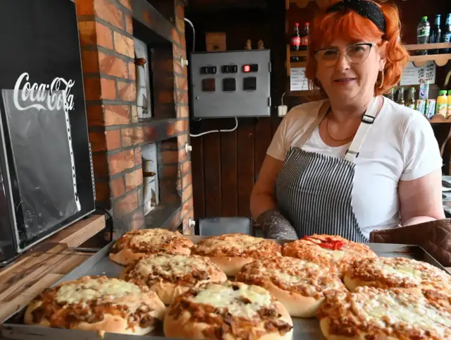 Kobieta z rudymi włosami prezentuje świeżo upieczone pizze. To może być najstarsza pizzeria w Polsce, z piecem opalanym drewnem w tle.
