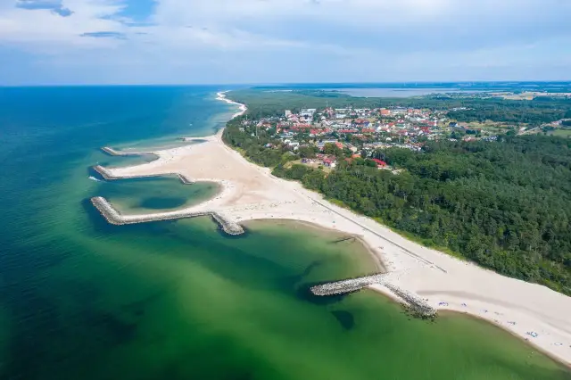 Widok z lotu ptaka na kładno nad morzem, z piaszczystą plażą, kamiennymi falochronami i zielonymi lasami otaczającymi miasteczko.