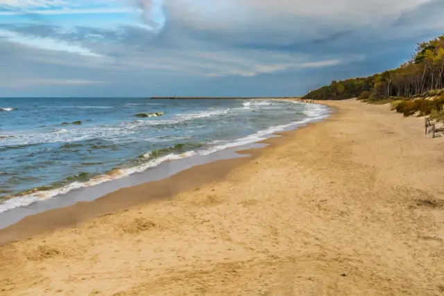 Plaża w Lubiatowie z piaszczystym brzegiem, falującym morzem i lasem w tle. W oddali widać falochron i latarnię morską.